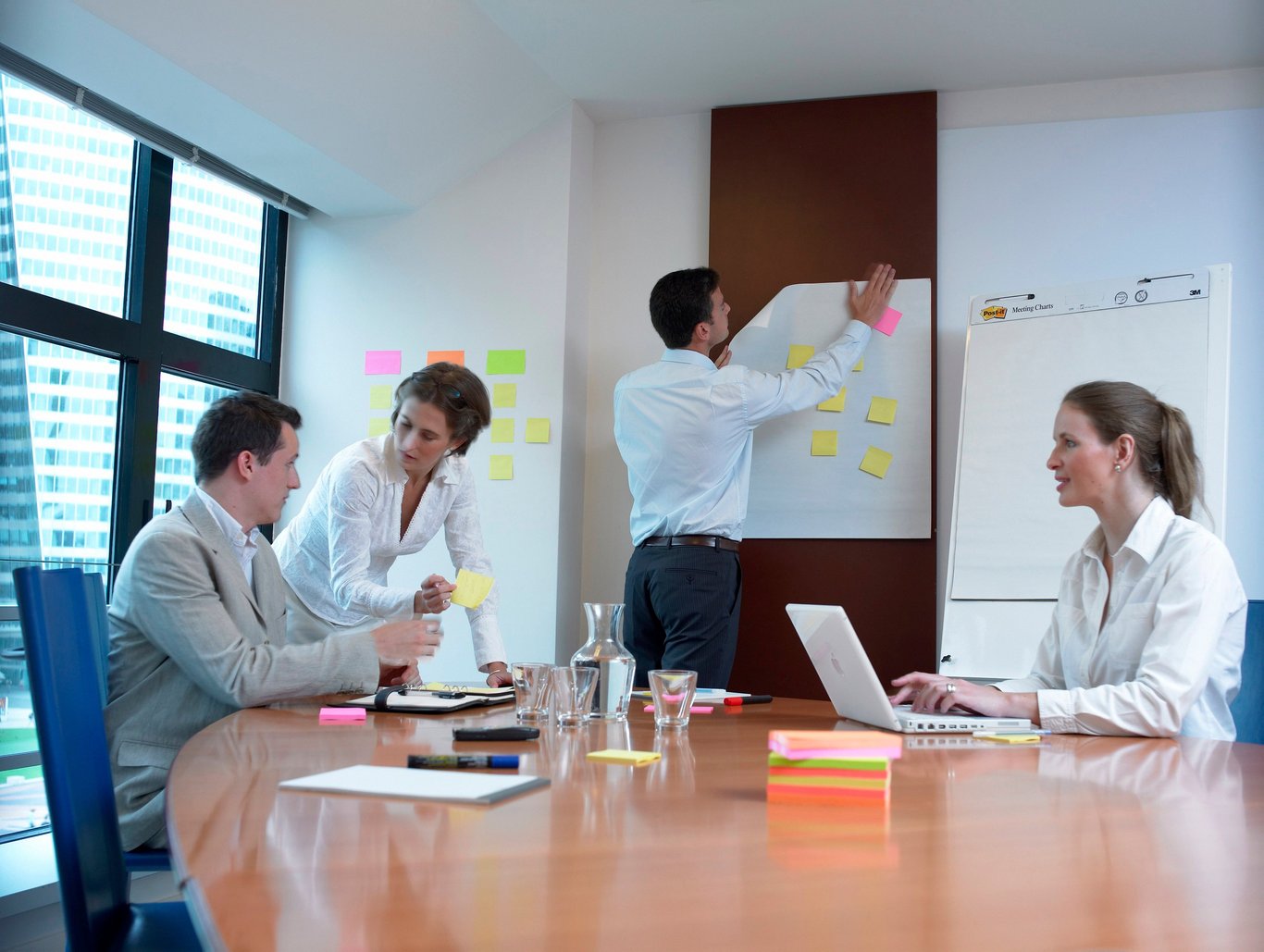 Mötesrum med tre personer, en skriver på blädderblock, andra två sitter vid bord med post-it lappar och laptop