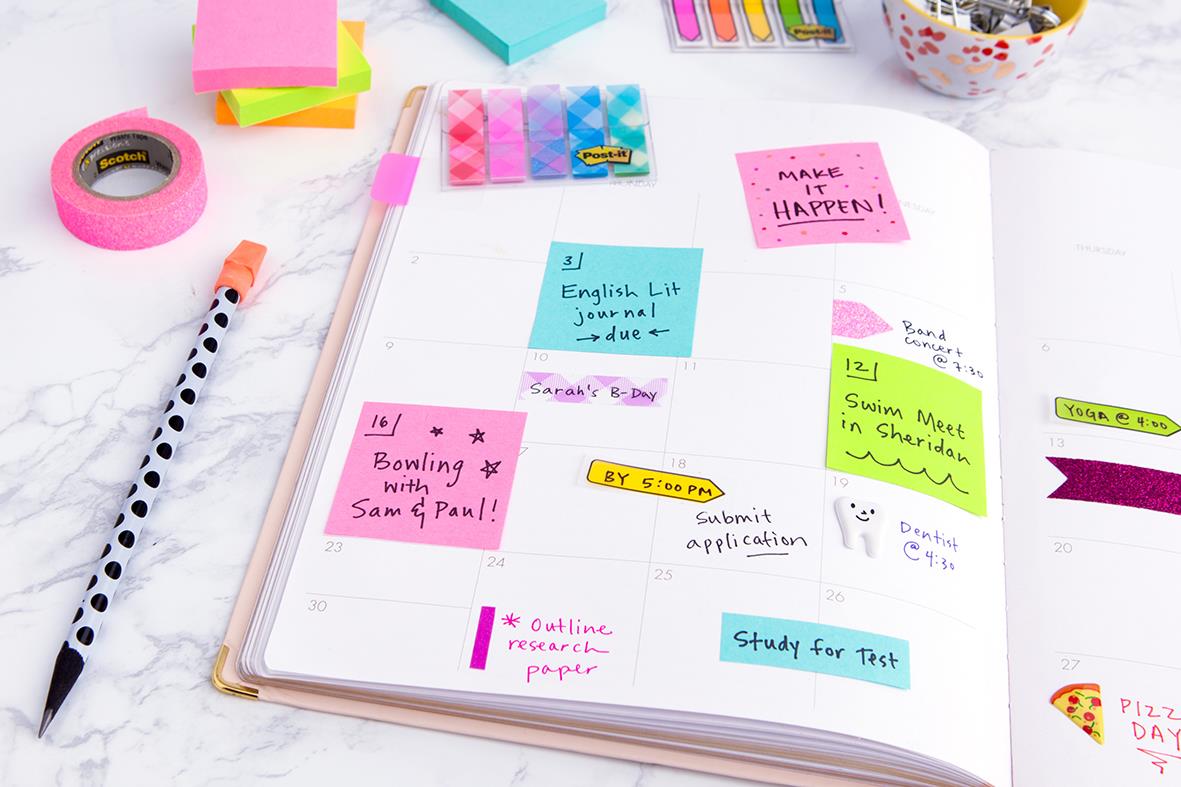 Post-it lappar i rosa, blå och gröna färger på en öppen anteckningsbok med skrivmaterial runtomkring