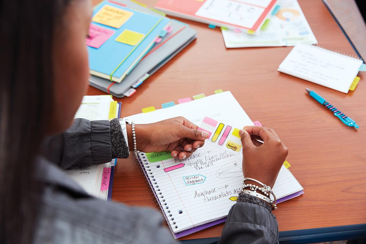 Person använder en hållare för Post-it-lappar i gult, svart och transparent på ett skrivbord med anteckningar och papper