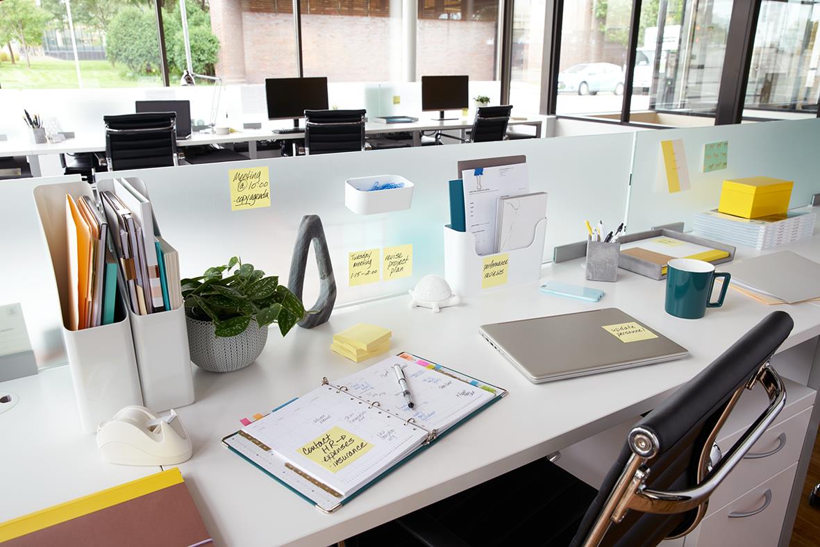 Kontorsbord med Post-it lappar, anteckningsblock, växt och kontorsmaterial i ett modernt kontorslandskap