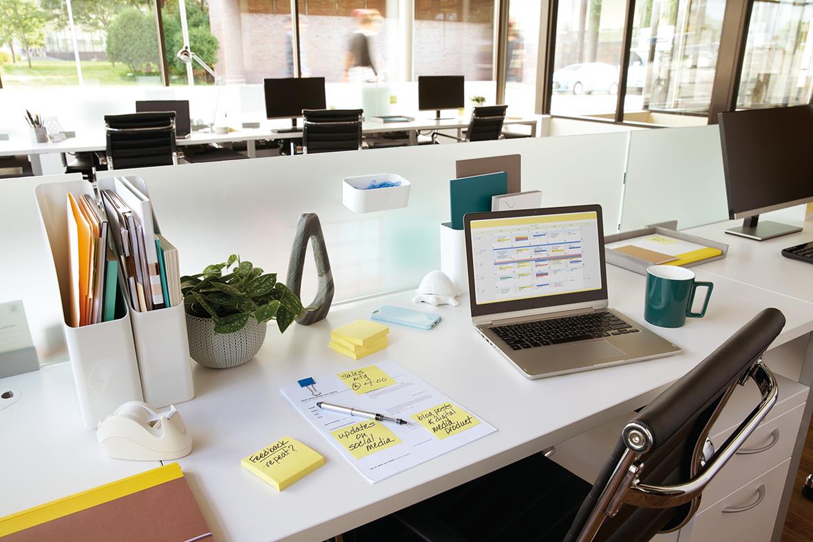 Kontorsbord med Post-it-gul lappar, laptop, muggar och kontorsmaterial i ett modernt kontorslandskap