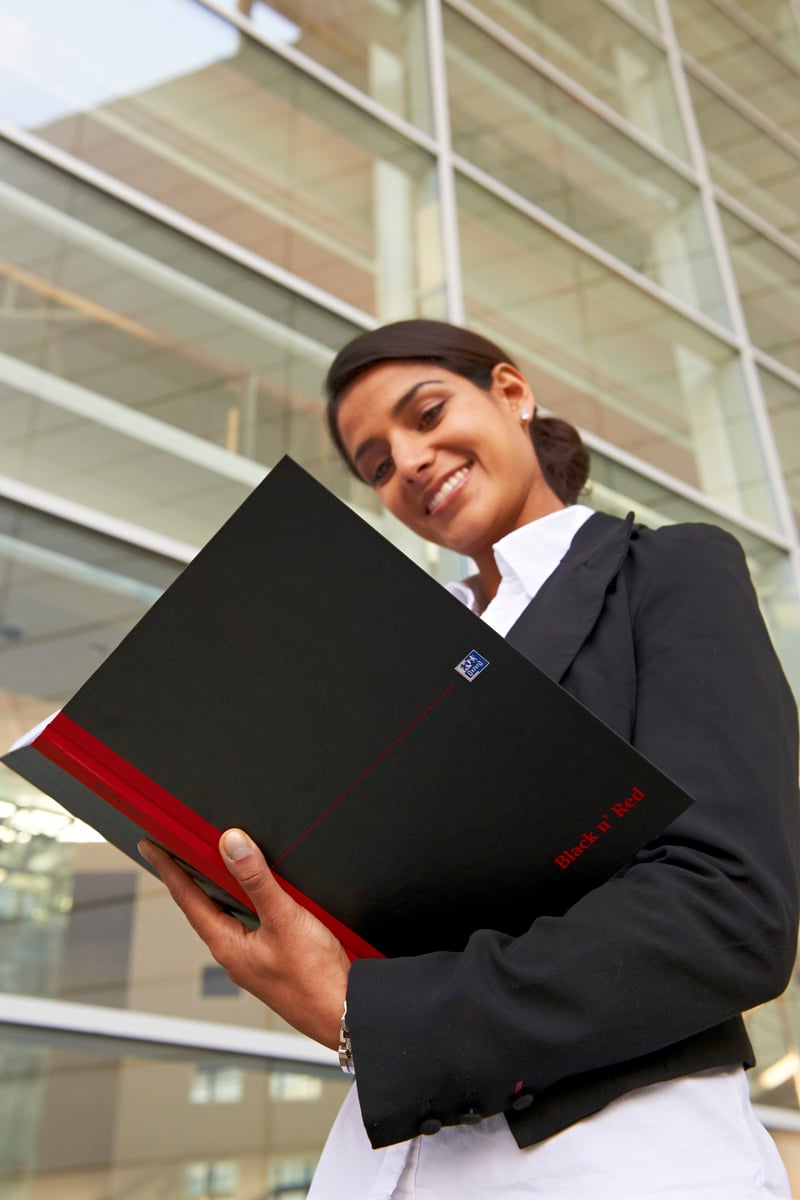 Kvinna i svart kavaj håller anteckningsbok Oxford Black n' Red Linjerad Svart A4 framför modern glasbyggnad