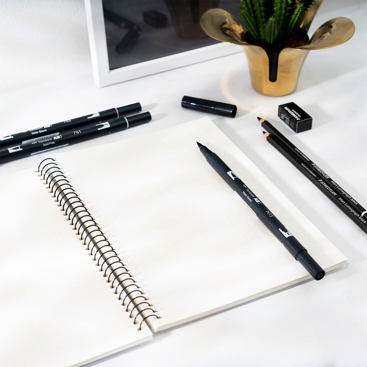 Öppen spiralanteckningsbok med svarta pennor och växt på skrivbordet