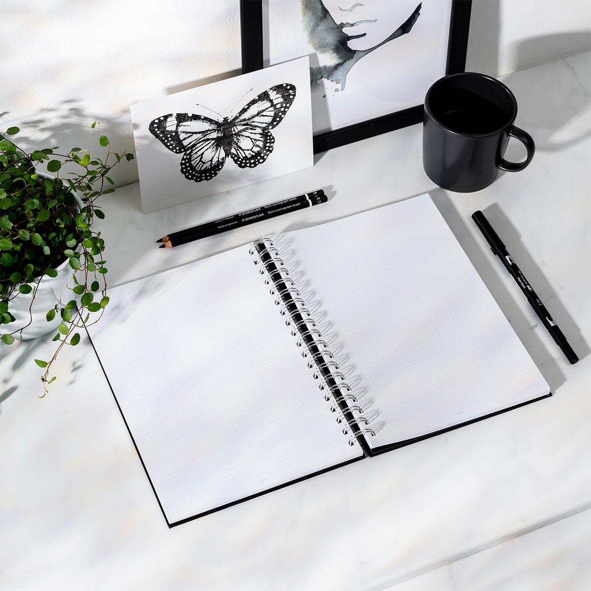 Svart anteckningsbok med spiralbindning, tomma sidor, omgiven av svart penna, svart kopp, växt och konstverk på skrivbordet