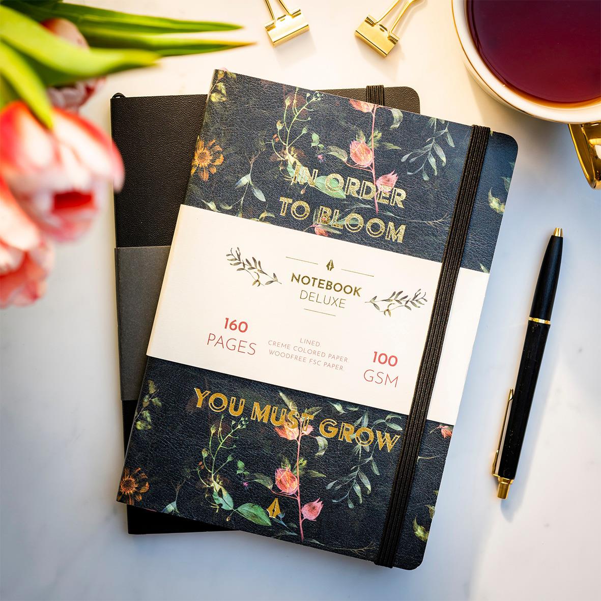 Anteckningsbok Burde Deluxe Flower A5 med blommönster, 160 sidor, omslag och elastisk band, placerad på vit yta med penna och kopp te.