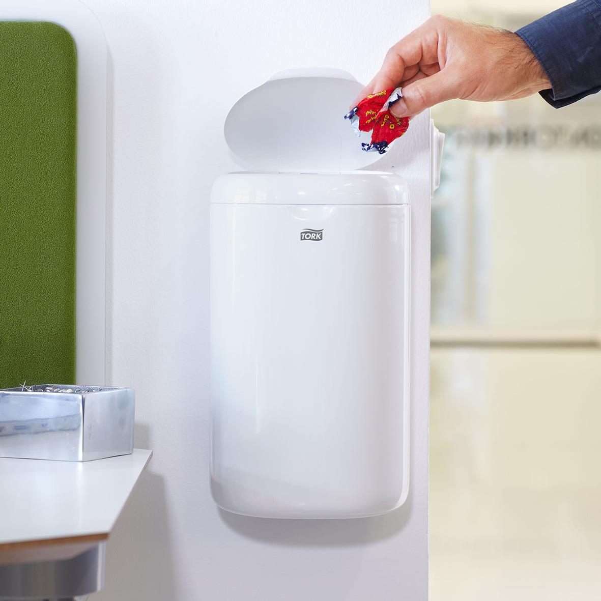 Person slänger skräp i vit papperskorg Tork B3 med lock, väggmonterad, 5 liter