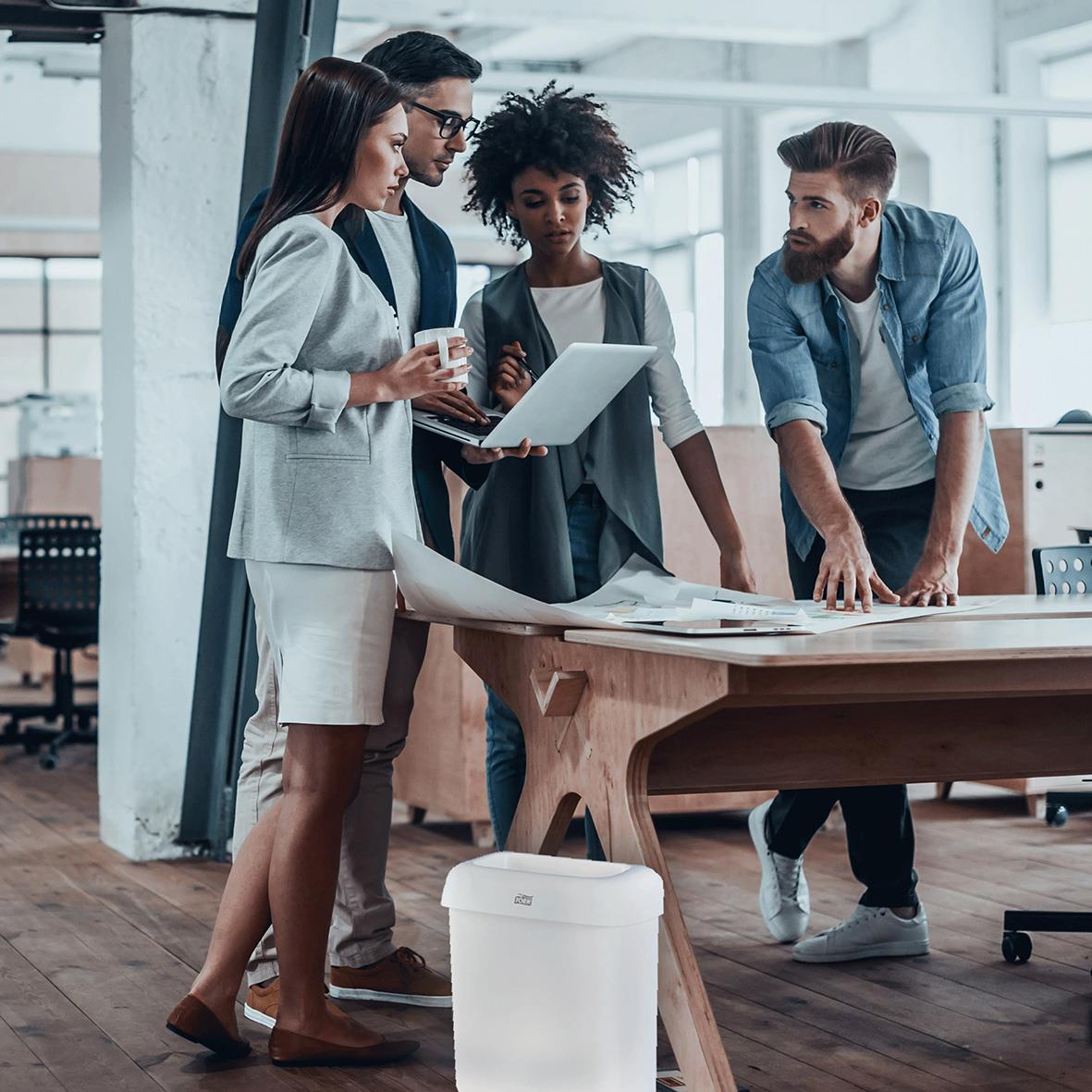 Fyra personer samlas runt ett bord i ett kontorslandskap, med en vit papperskorg Tork B2 Plast Vit 20L på golvet.