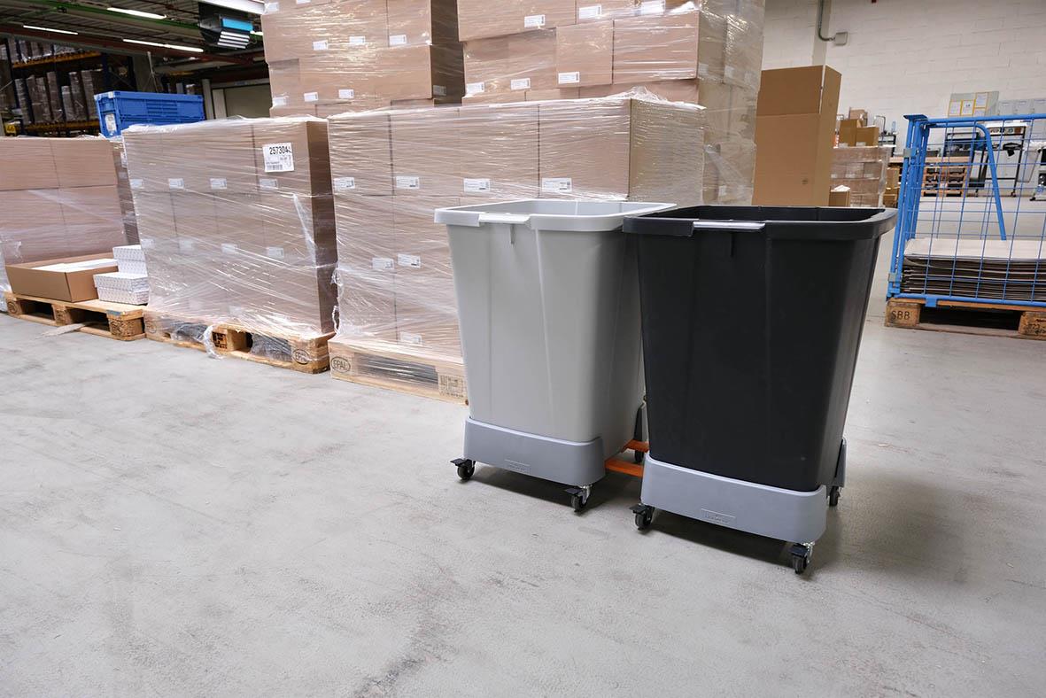 Två sopkärl i olika färger står i ett lagerutrymme med pallar och kartonger i bakgrunden