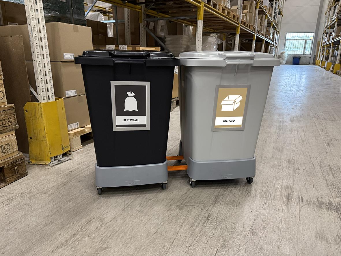 Två sopkärl för avfallshantering i ett lager, en svart och en grå, med symboler för restavfall och förpackningar.