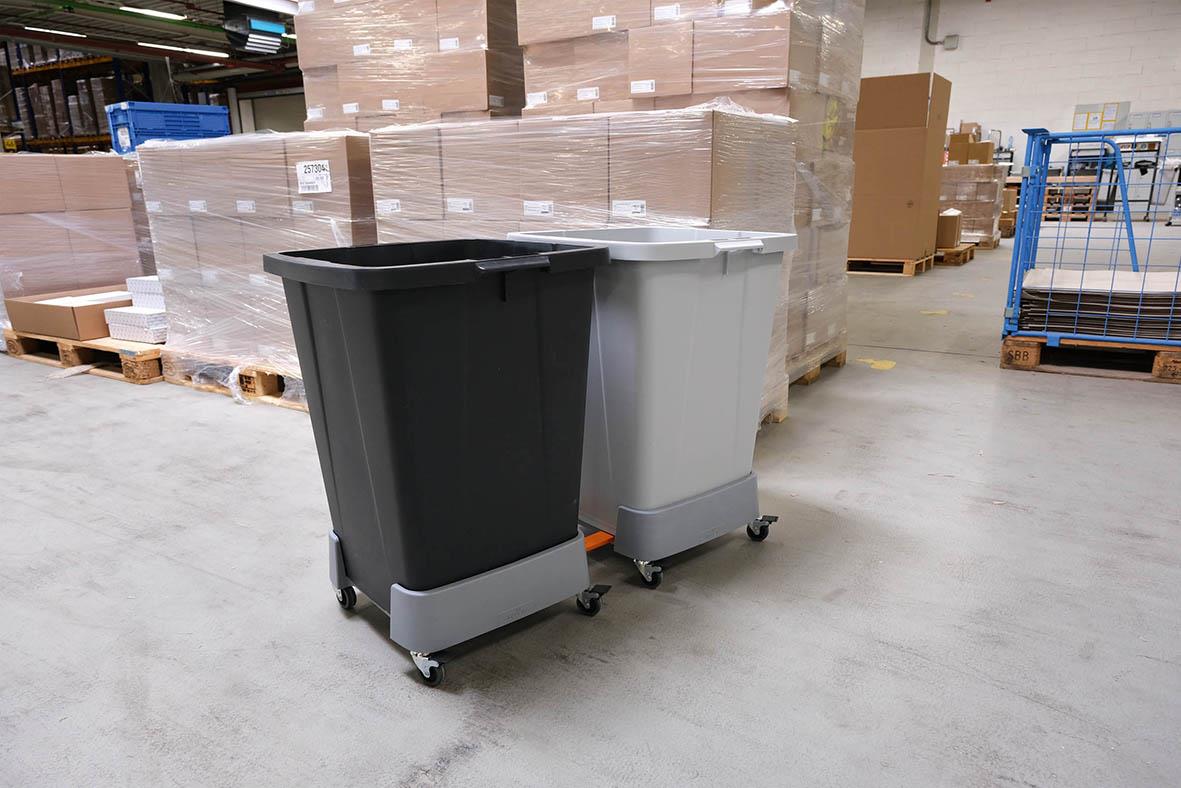 Två stora sopkärl, en svart och en grå, på hjul i lagerlokal med pallar och förpackningar i bakgrunden