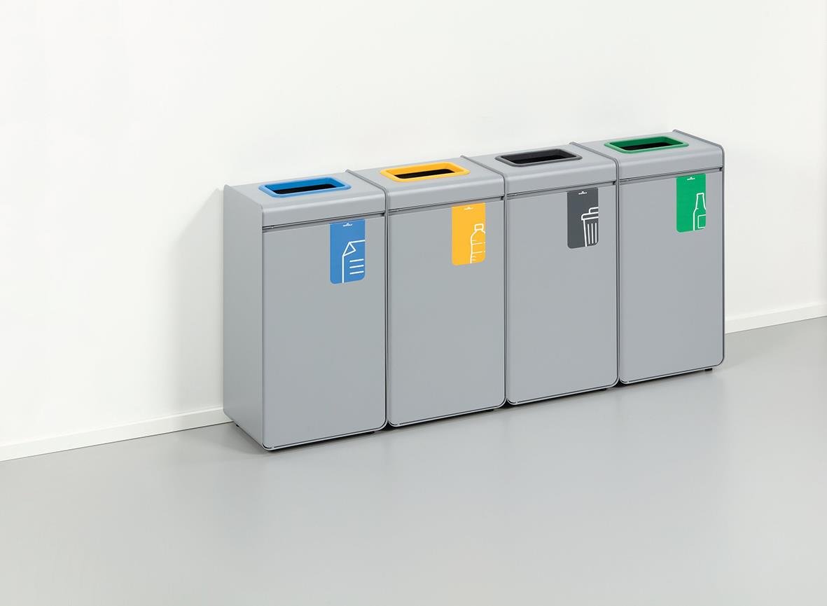 Fyra återvinningskärl i grått med färgkodade lock för papper, plast, metall och glas, placerade mot en vit vägg
