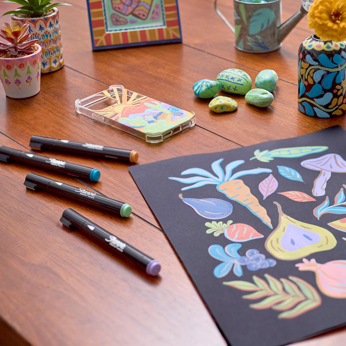Skissblock med färgglada växt- och fruktillustrationer, pennor, stenar och blomkrukor på ett träbord