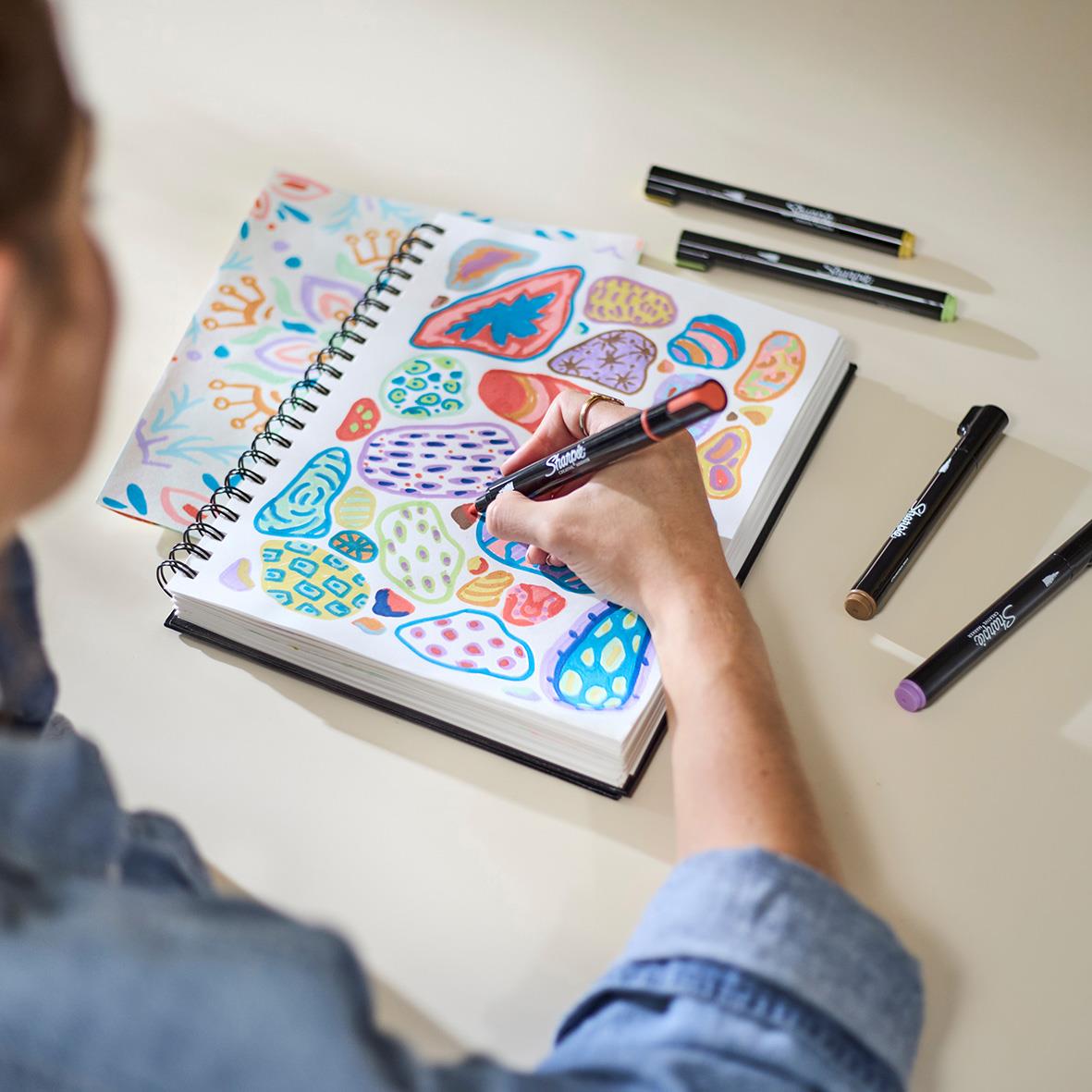 Person som ritar med en Sharpie Creative Earth Tones pen på en skissbok med färgglada mönster