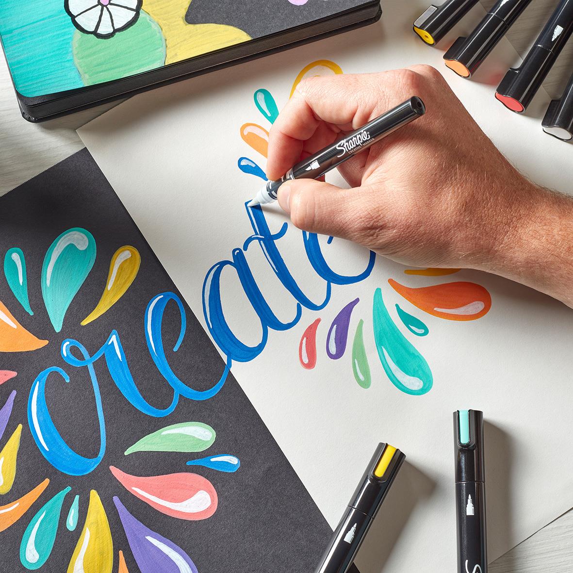 Hand som ritar med Sharpie Creative markers i olika färger på papper med färgglada design och markörer runt omkring