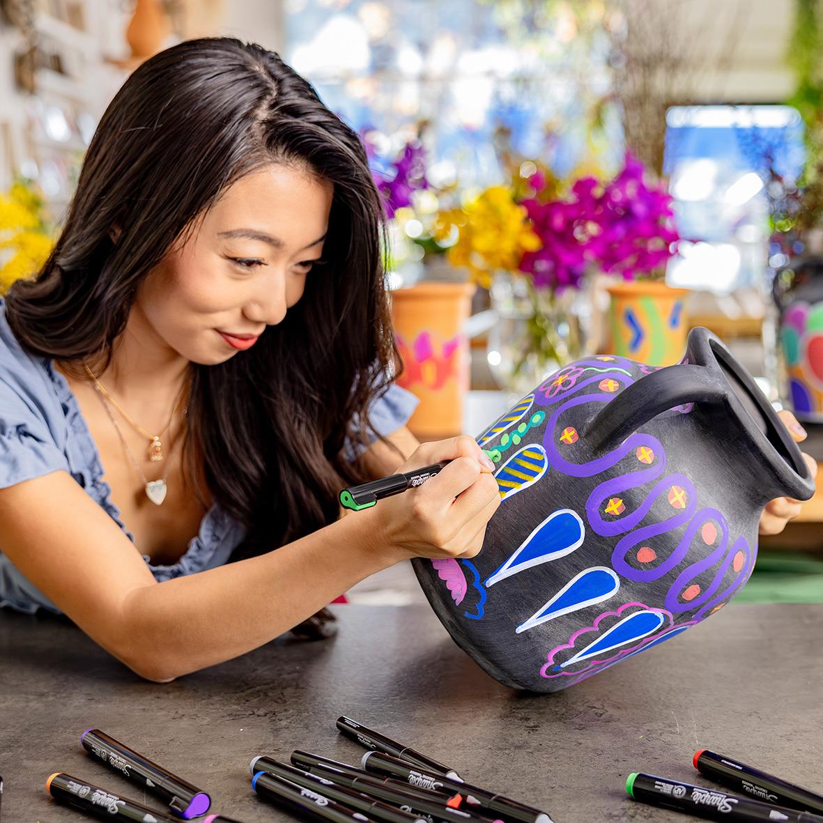 Kvinna målar ett svart krus med färgglada Sharpie Creative pennor med penselpipett, omgiven av blommor och konstmaterial