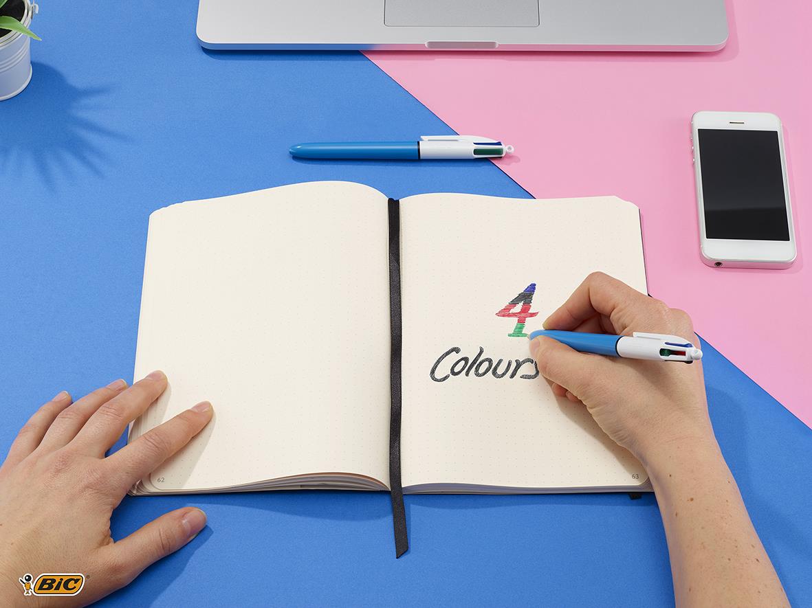 Person skriver med en 4-färg kulspetspenna i ett öppet anteckningsblock på blå och rosa bakgrund