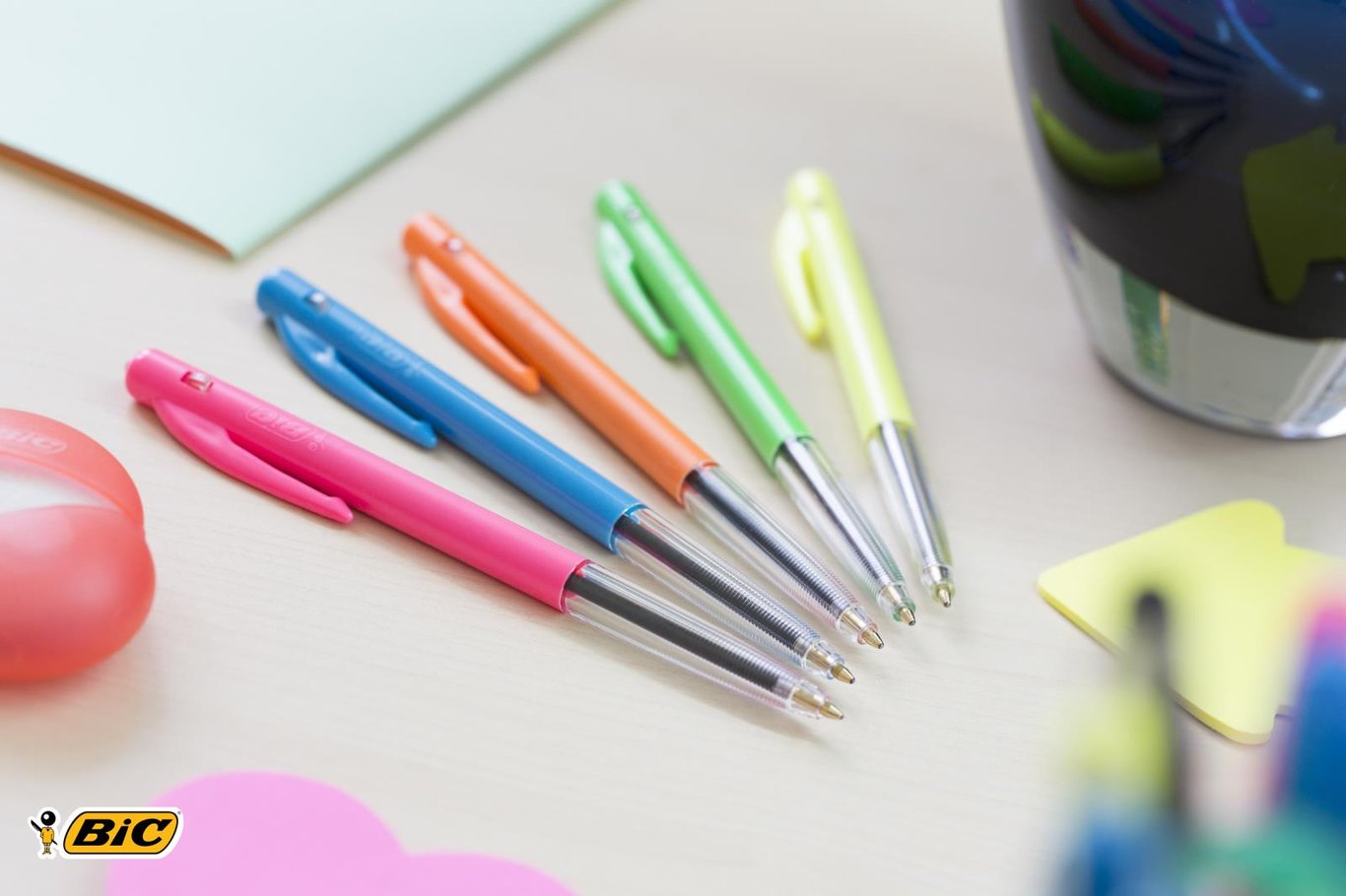 Fem kulspetspennor i olika färger på ett skrivbord med andra kontorsmaterial