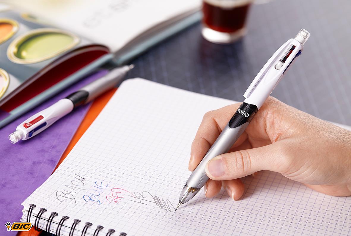 Hand som skriver med en kulspetspenna Bic på ett rutat papper, omgiven av andra pennor och skrivmaterial
