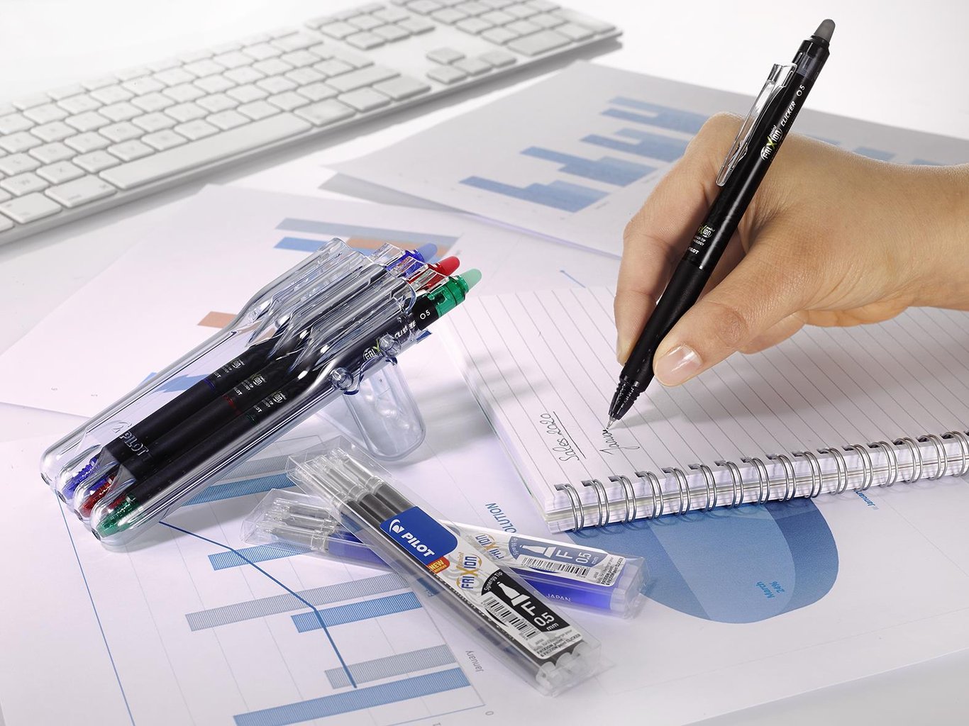 Hand som skriver med en Pilot Frixion Point Clicker 4-färg penna på papper med diagram och dator i bakgrunden