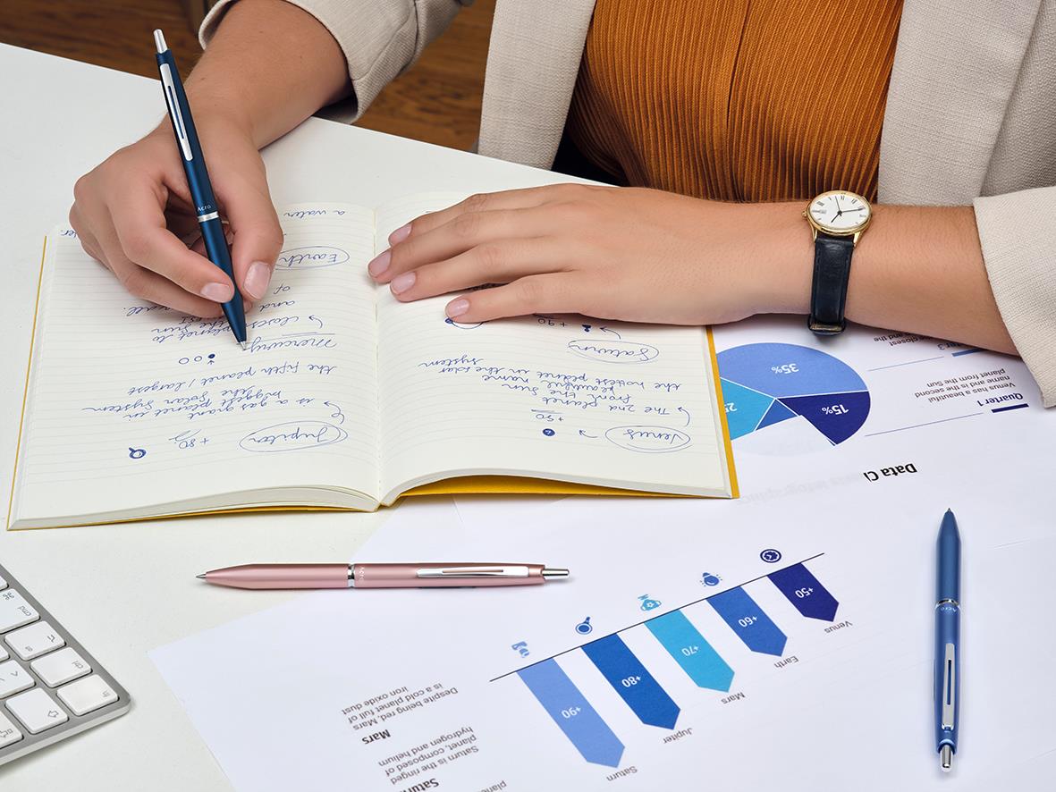Person skriver i anteckningsbok med penna, omgiven av diagram och papper på skrivbordet