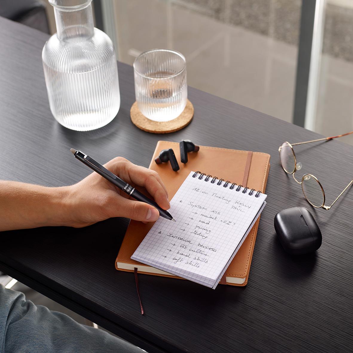 Person skriver i ett anteckningsblock på ett svart bord med glas, hörlurar och en penna