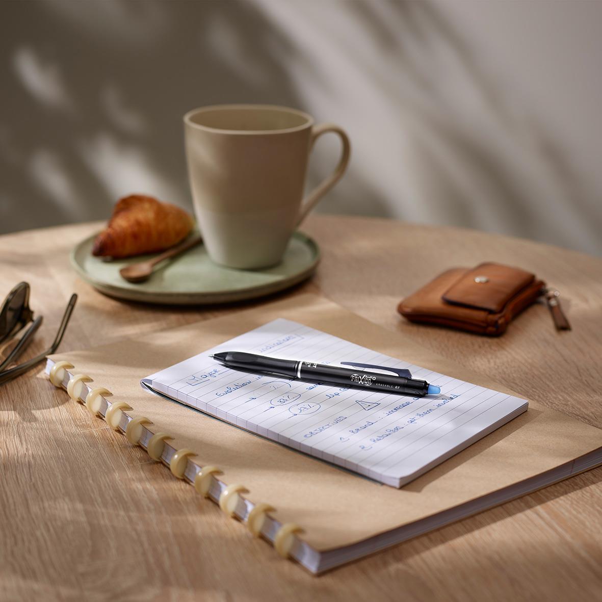 Skrivblock, penna, kopp, croissant och plånbok på ett bord i ett mysigt arbetsrum