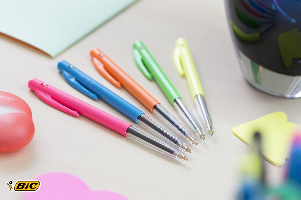 Fem kulspetspennor i pastellfärger rosa blå orange grön och gul på skrivbord med andra kontorsmaterial