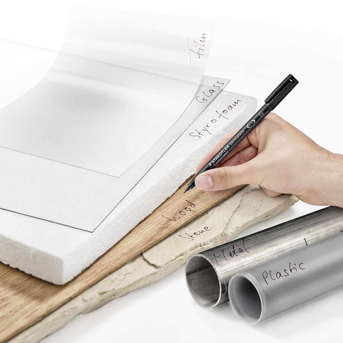 Hand som skriver med en svart permanent penna på papper, omgiven av olika material som plaströr och trä.
