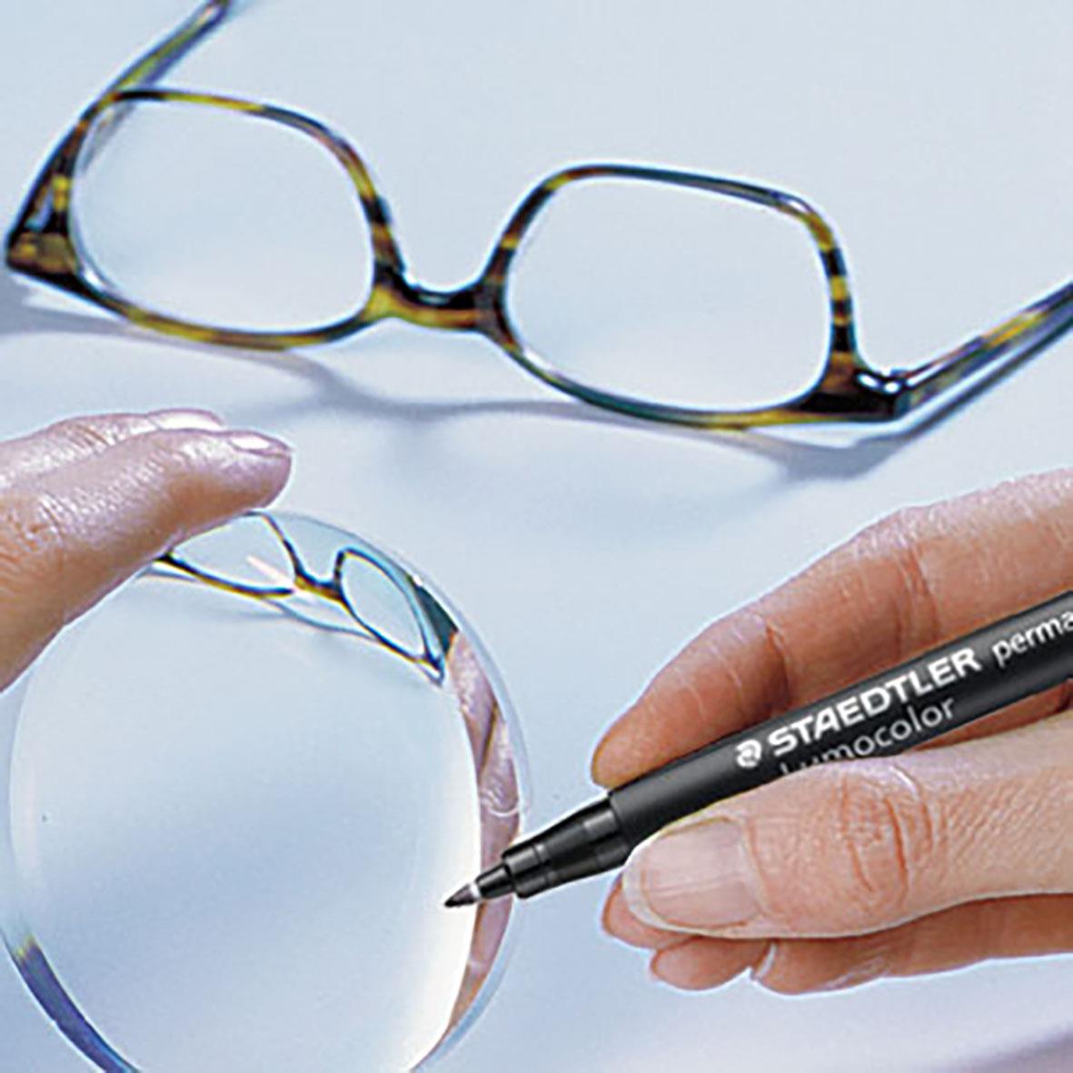 Närbild av en svart Staedtler Lumocolor 319 permanent penna som används för att skriva på ett glas