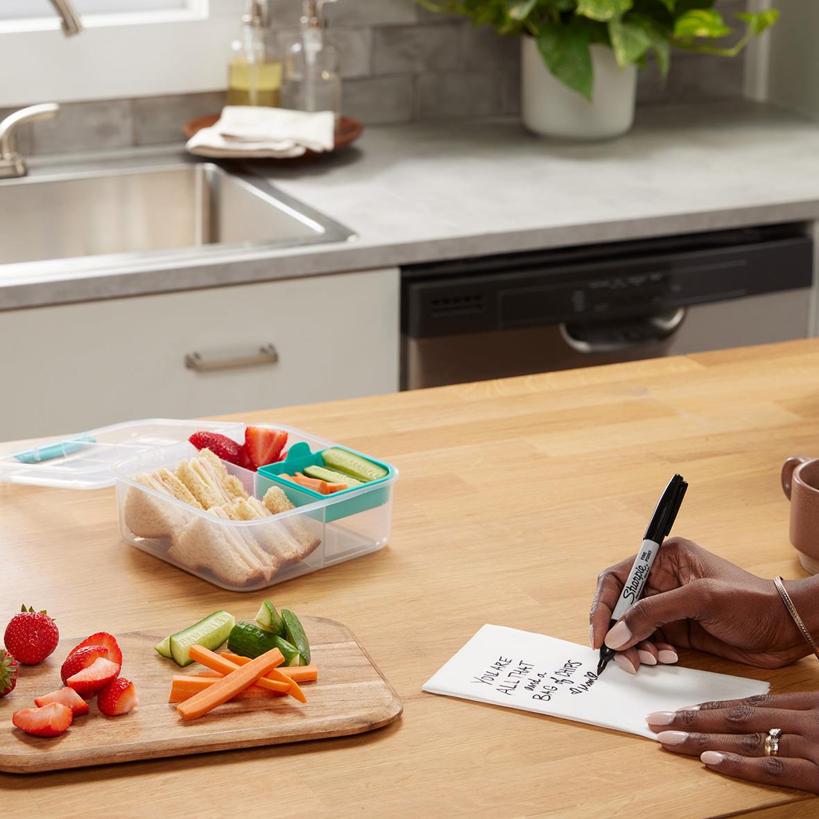 Person skriver på en liten whiteboard eller papper med en svart permanent marker i ett kök med träbänk och frukt på skärbräda