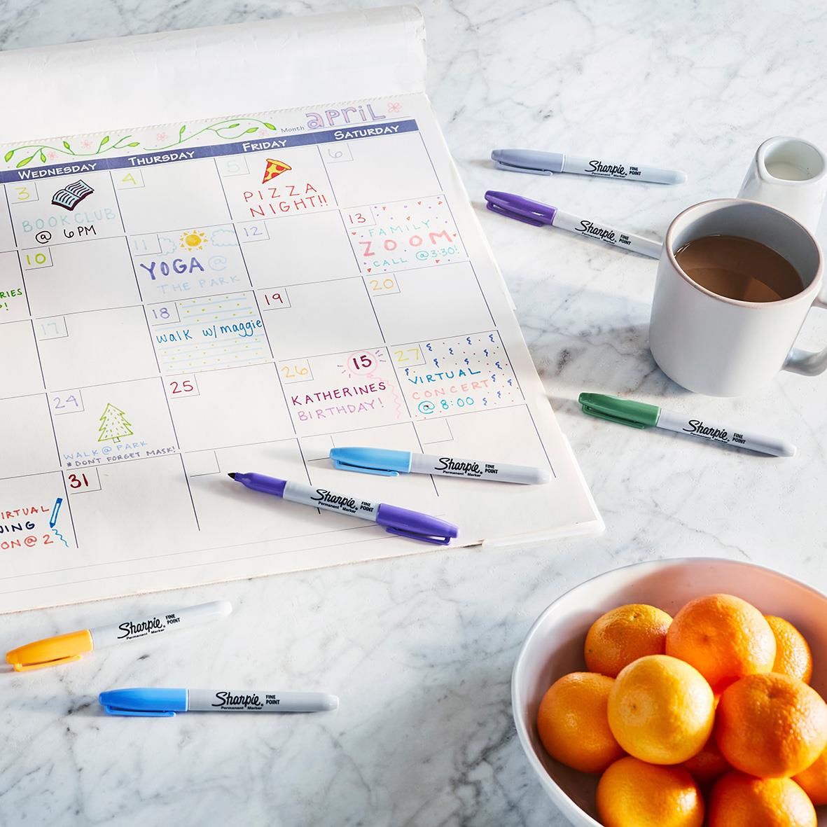 Kalender med färgade pennor, kopp kaffe och en skål med små apelsiner på ett vitt bord