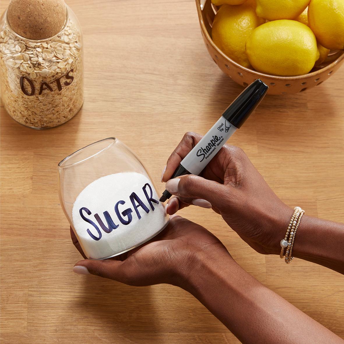 Person skriver SUGAR på ett glas med svart permanent marker, omgiven av frukt och köksartiklar