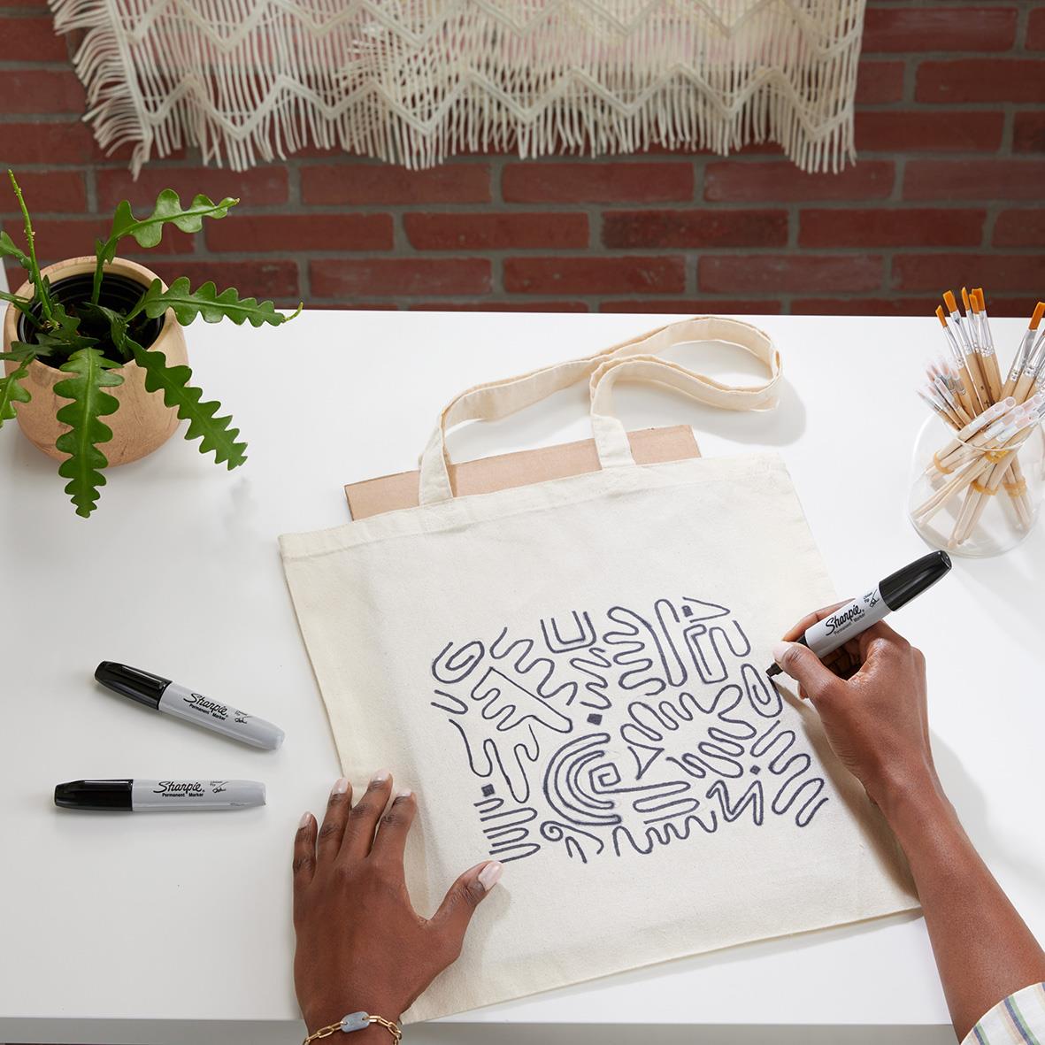 Person skriver på en tygpåse med svart permanent marker, omgiven av växter och skrivmaterial