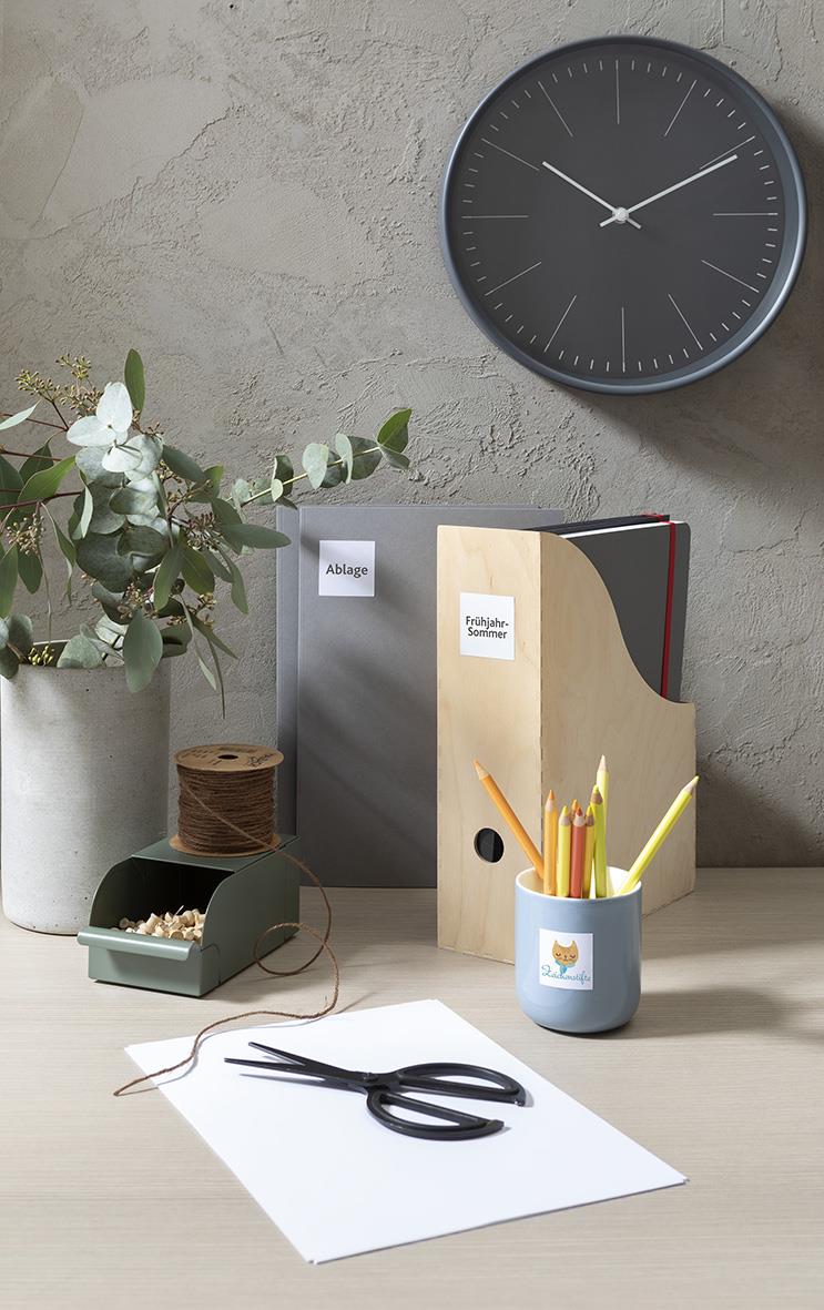 Väggklocka på grå vägg, skrivbord med papper, sax, pennor, blomkruka med gröna blad, filofax och etikettark