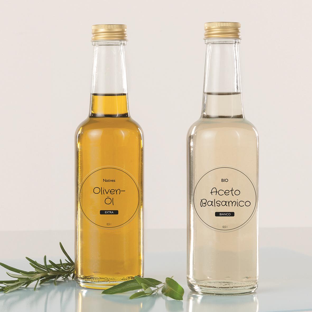Två transparenta glasflaskor med guld- och silverlock, innehållande olja och vinäger, med gröna örter bredvid