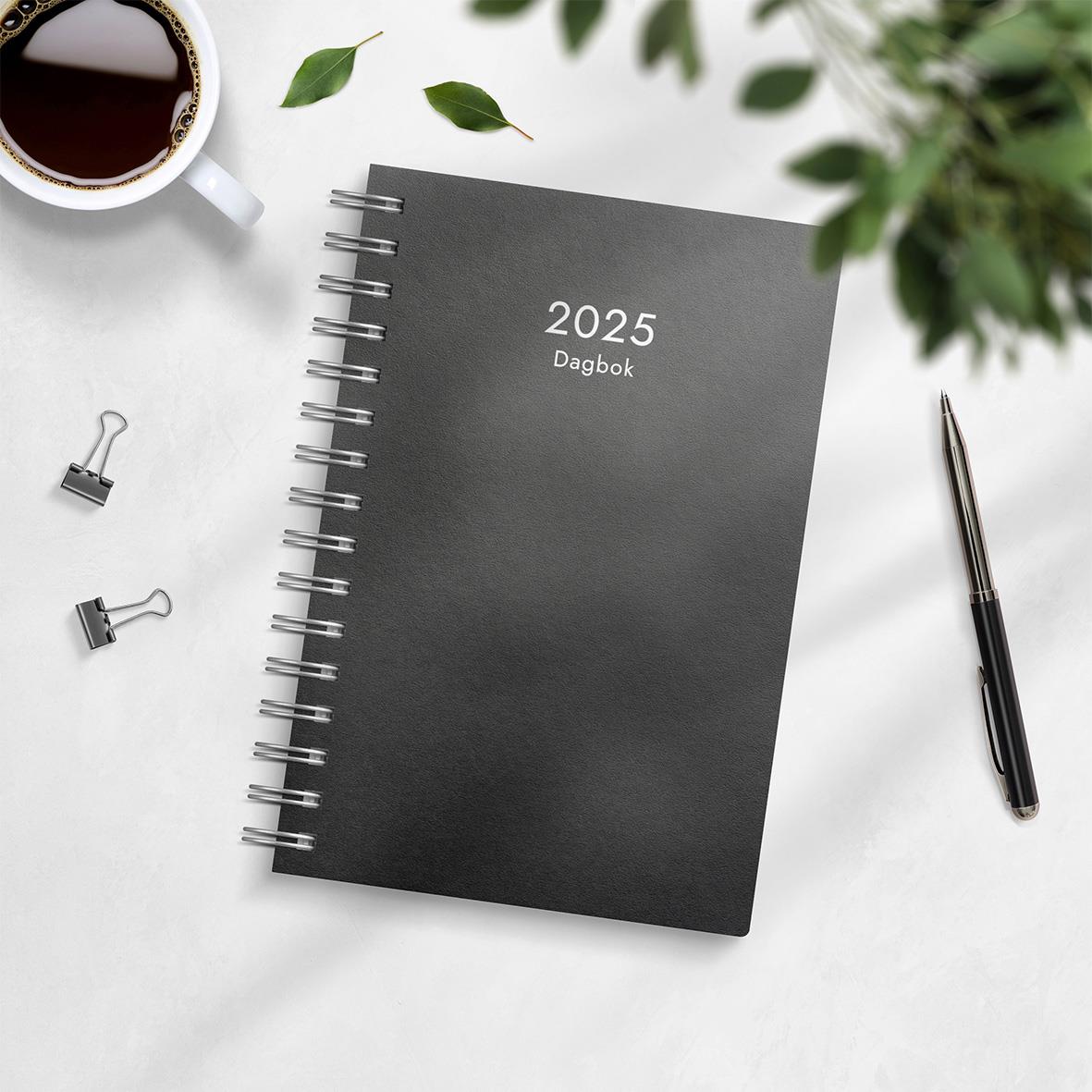 Svart dagboksrefill för 2026 med spiralbindning, omgiven av penna, klämmor, och en kopp kaffe på ett vitt bord