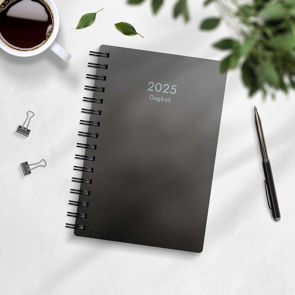 Svart 2025 dagbok med spiralbindning och PP-plastomslag, omgiven av bläckpenna, klämmor, kaffekopp och gröna växter