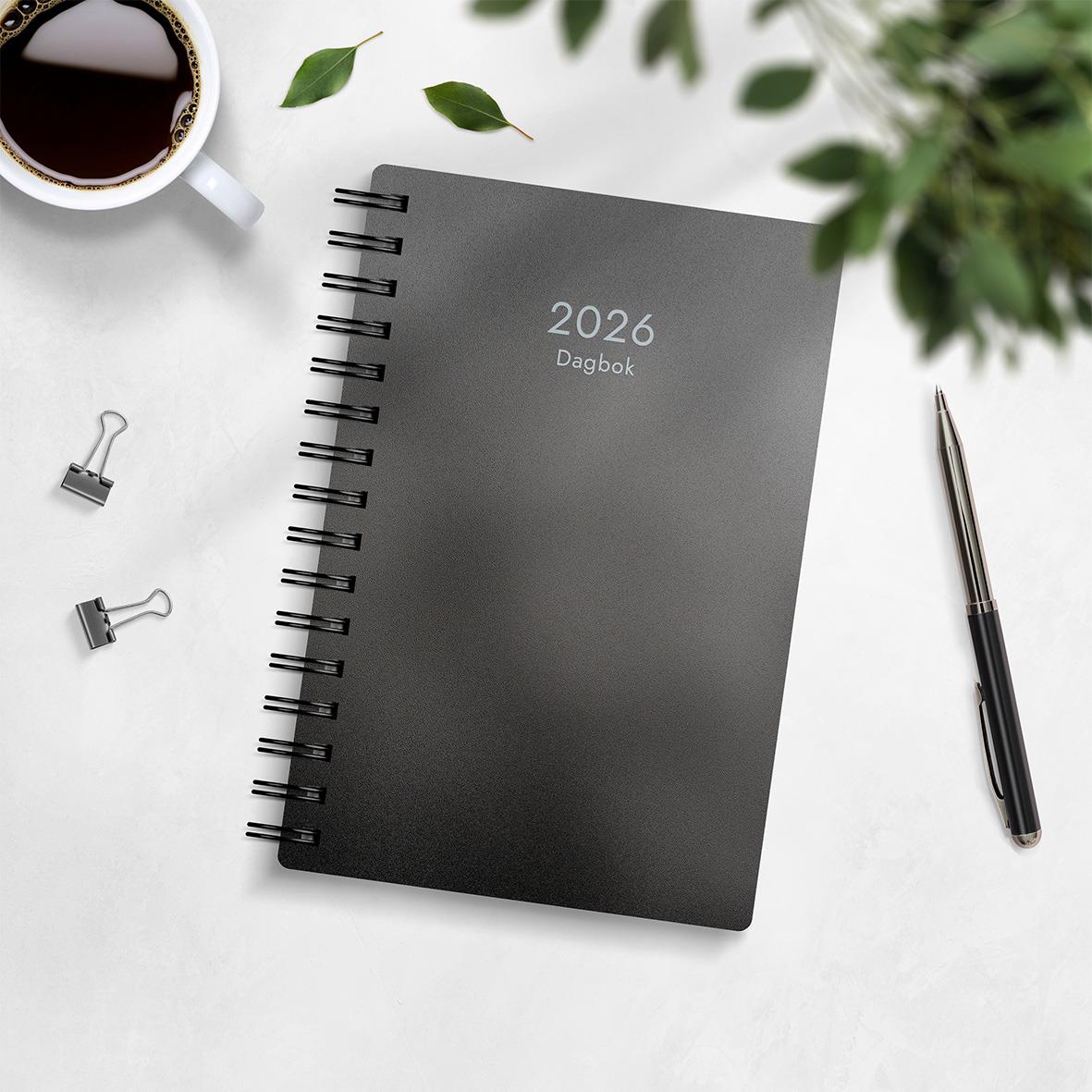 Svart 2026 dagbok i plastomslag med spiralbindning, omgiven av penna, klämmor, kaffekopp och gröna växter