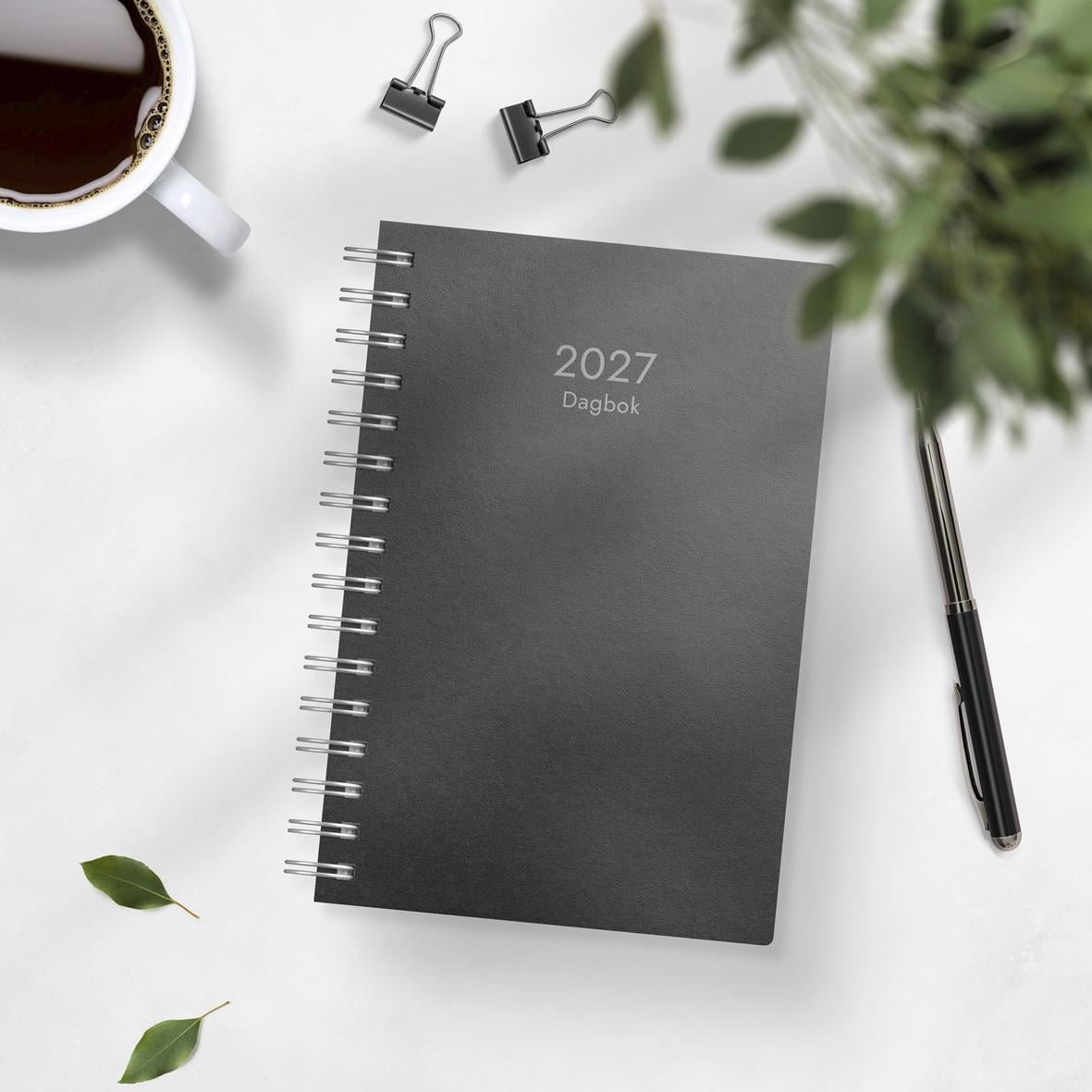 Svart 2025 dagbok med spiralbindning och penna på vit bakgrund med kaffekopp och gröna växter