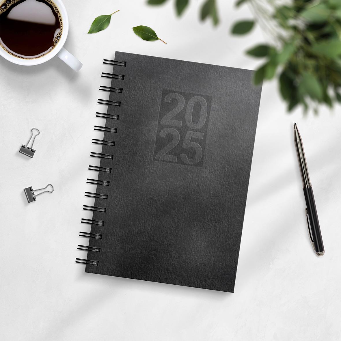 Svart 2025 års dagbok med spiralbindning och konstläderomslag, omgiven av penna, klämmor, kaffe och gröna växter