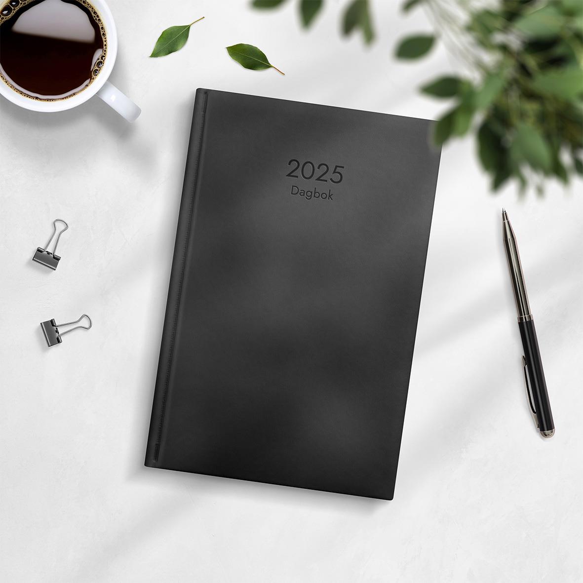 Svart 2025 dagbok i konstläder med inbindning och kalender för 2026, placerad på vit yta med penna, koppar och gröna blad