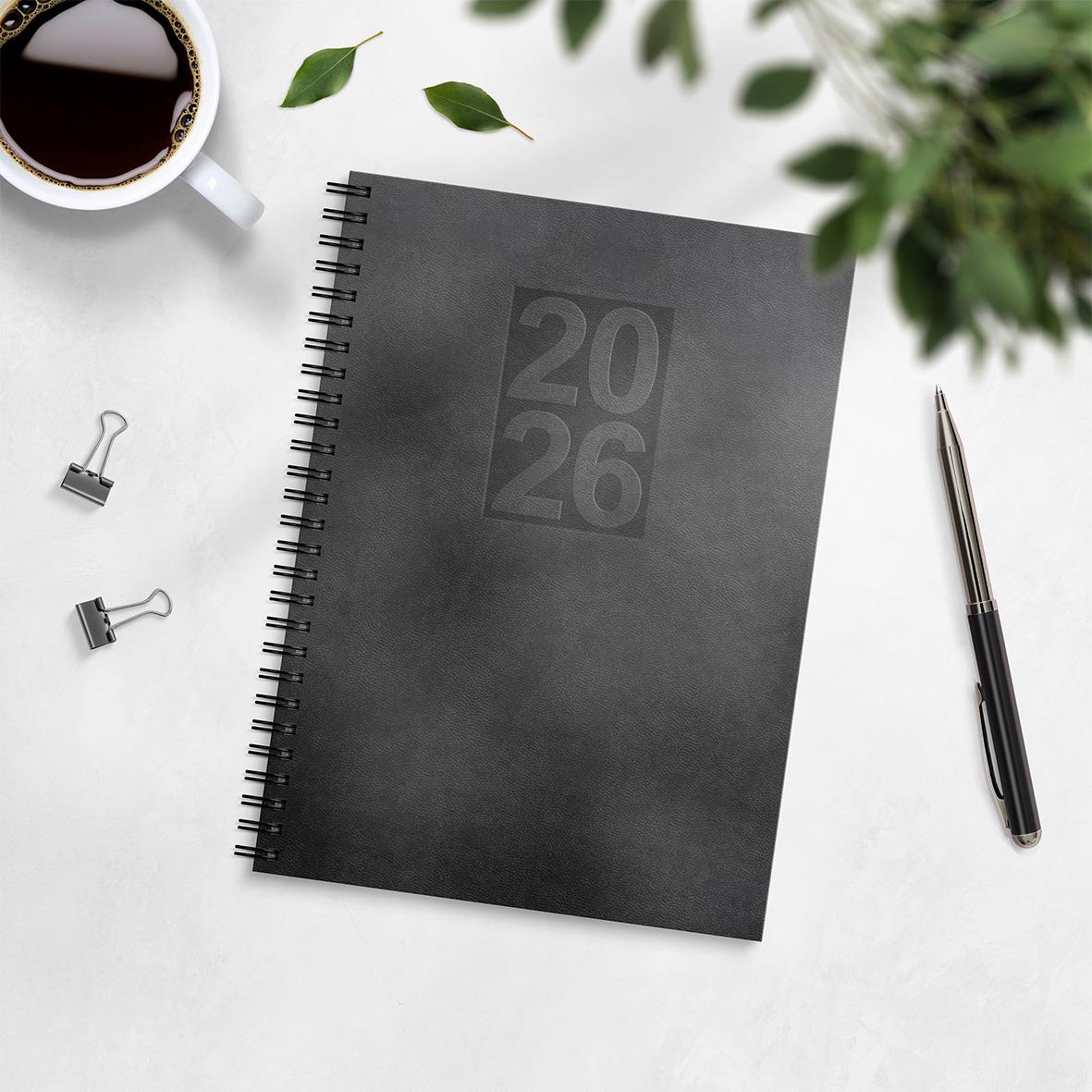 Svart almanacka för 2026 med konstläderomslag och spiralbindning på vit bord med kopp kaffe, penna och gröna växter