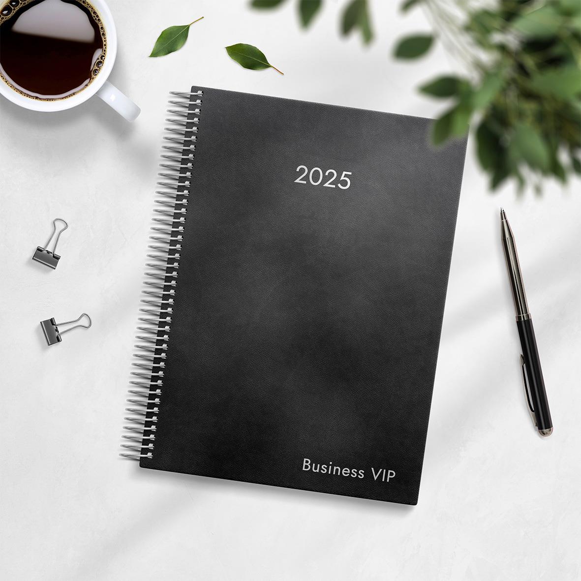 Svart affärsplanerare för 2025 med spiralbindning och text Business VIP på omslaget, omgiven av penna, klämmor, kaffekopp och gröna växter