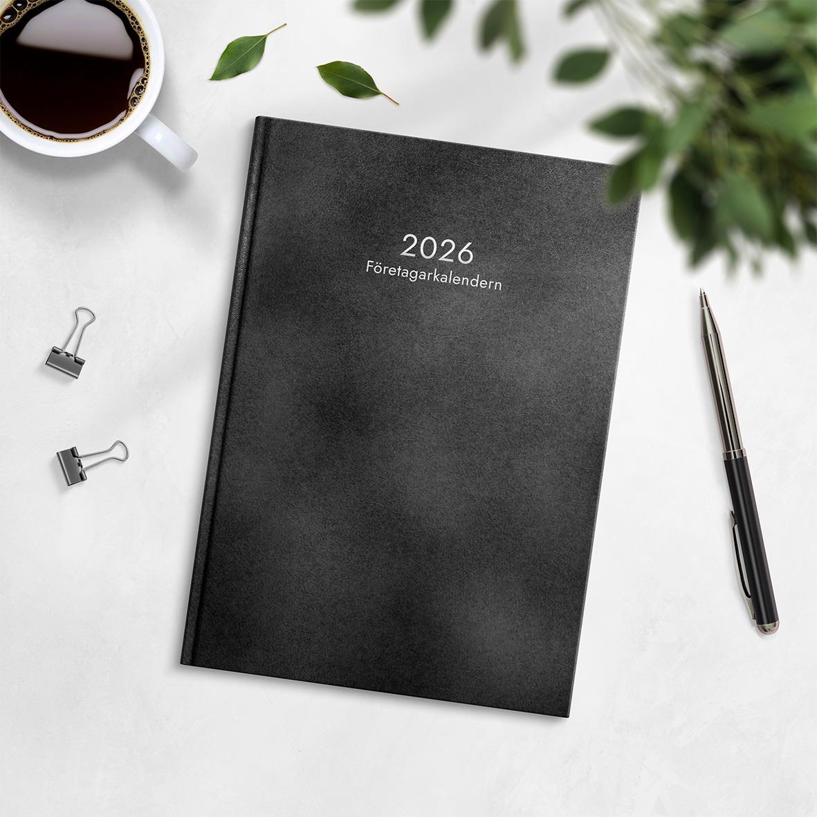 Företagarkalender 2026 i konstläder med svart omslag på vit skrivbord med kopp kaffe och kontorsmaterial