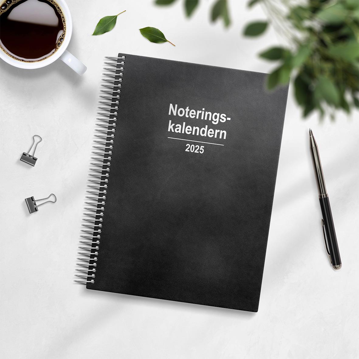 Svart noteringskalender för 2025 med spiralbindning och konstläderomslag, omgiven av penna, klämmor, kaffekopp och gröna växter