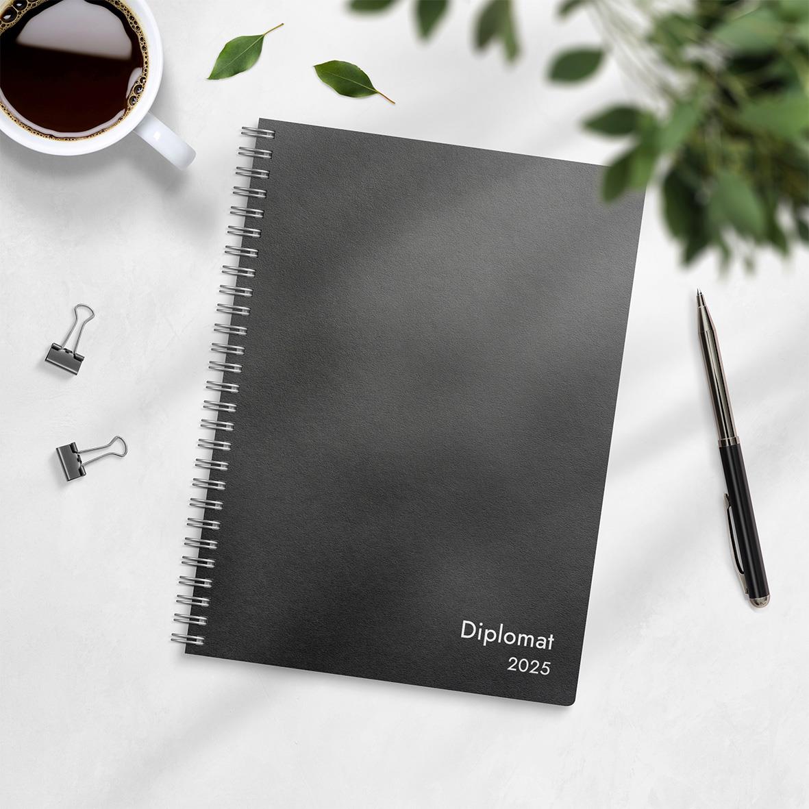 Svart spiralbunden almanacka Diplomat 2025 med pen, klämmor, kaffekopp och gröna blad runt