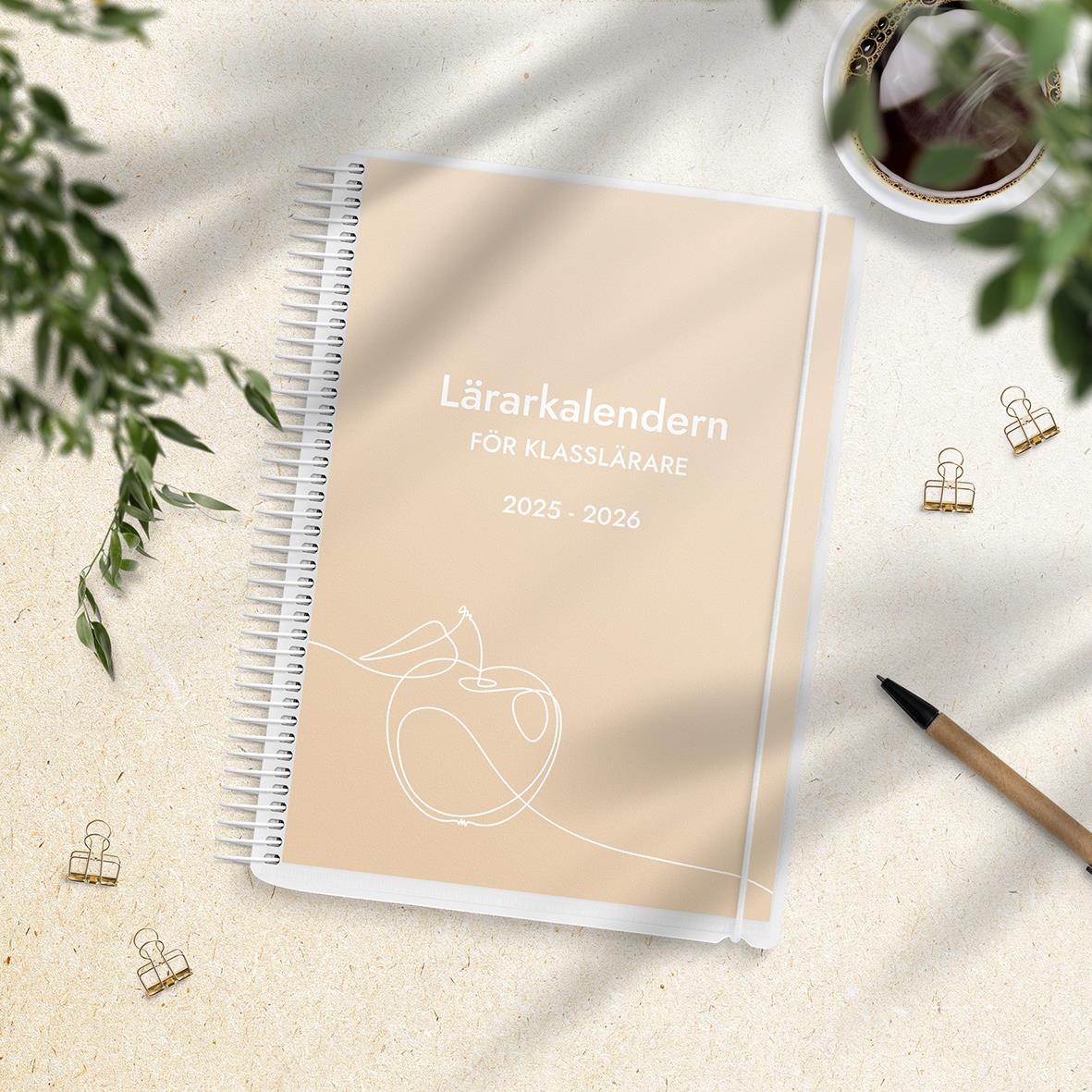 Lärarkalender för klasslärare 2025-2026 med spiralbindning, omgiven av gröna växter, penna och klämmor på ett vitt bord