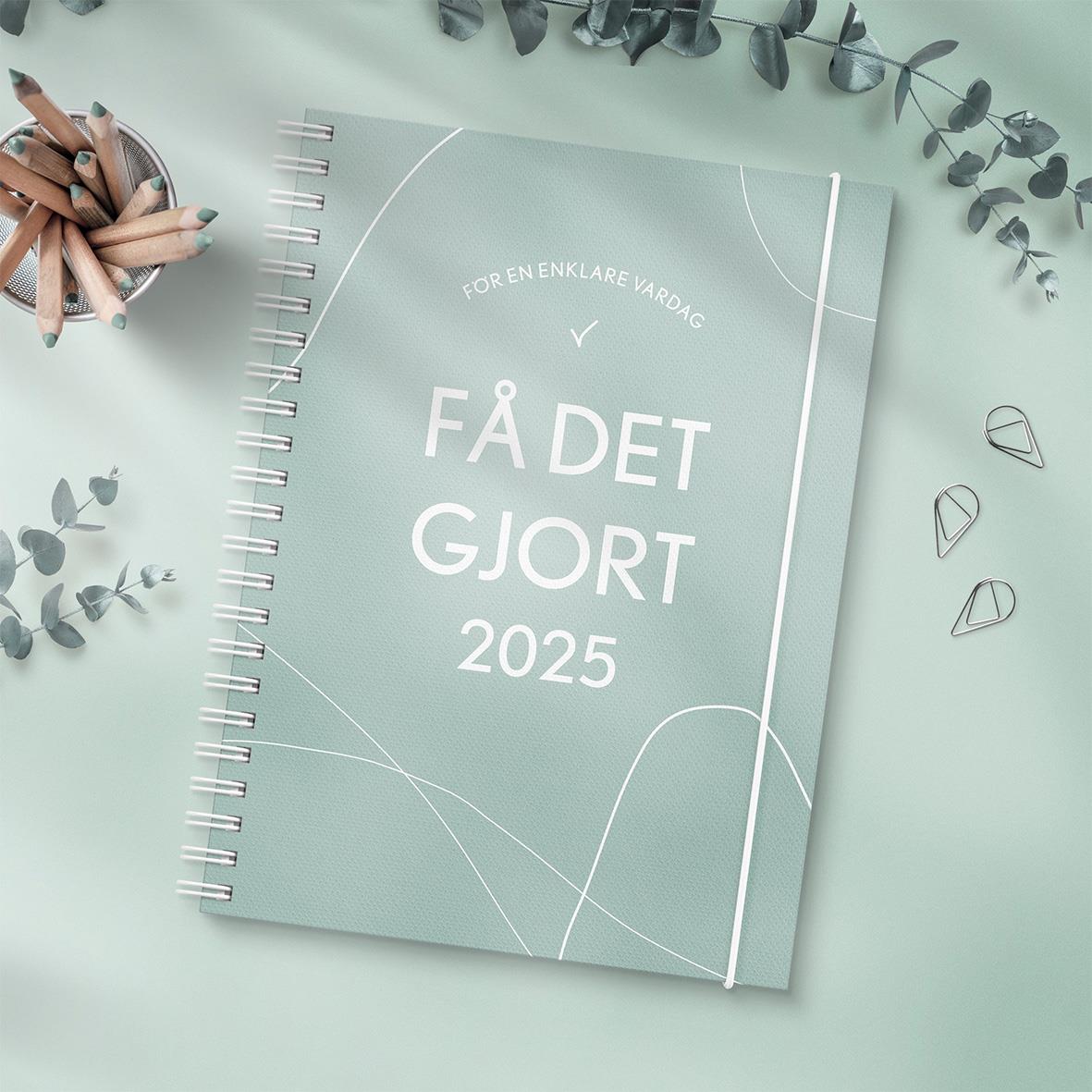 Grön A5 almanacka med texten Få det gjort 2025 och spiralrygg, omgiven av bläckpennor och växtdetaljer