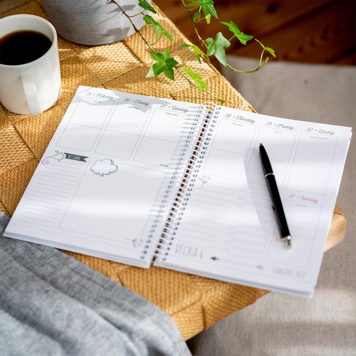 Öppen veckoplaneringsalmanacka med doodle för 2026, penna, kaffe och växt på träbord