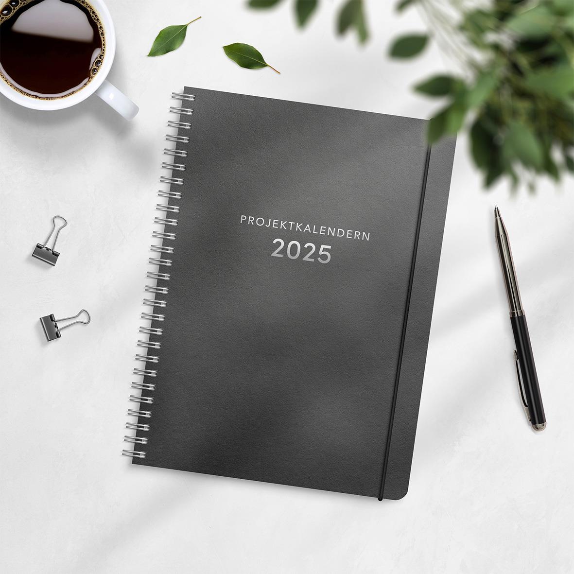 Projektkalender 2025 i svart omslag med spiralbindning, omgiven av penna, klämmor, kaffe och gröna växter